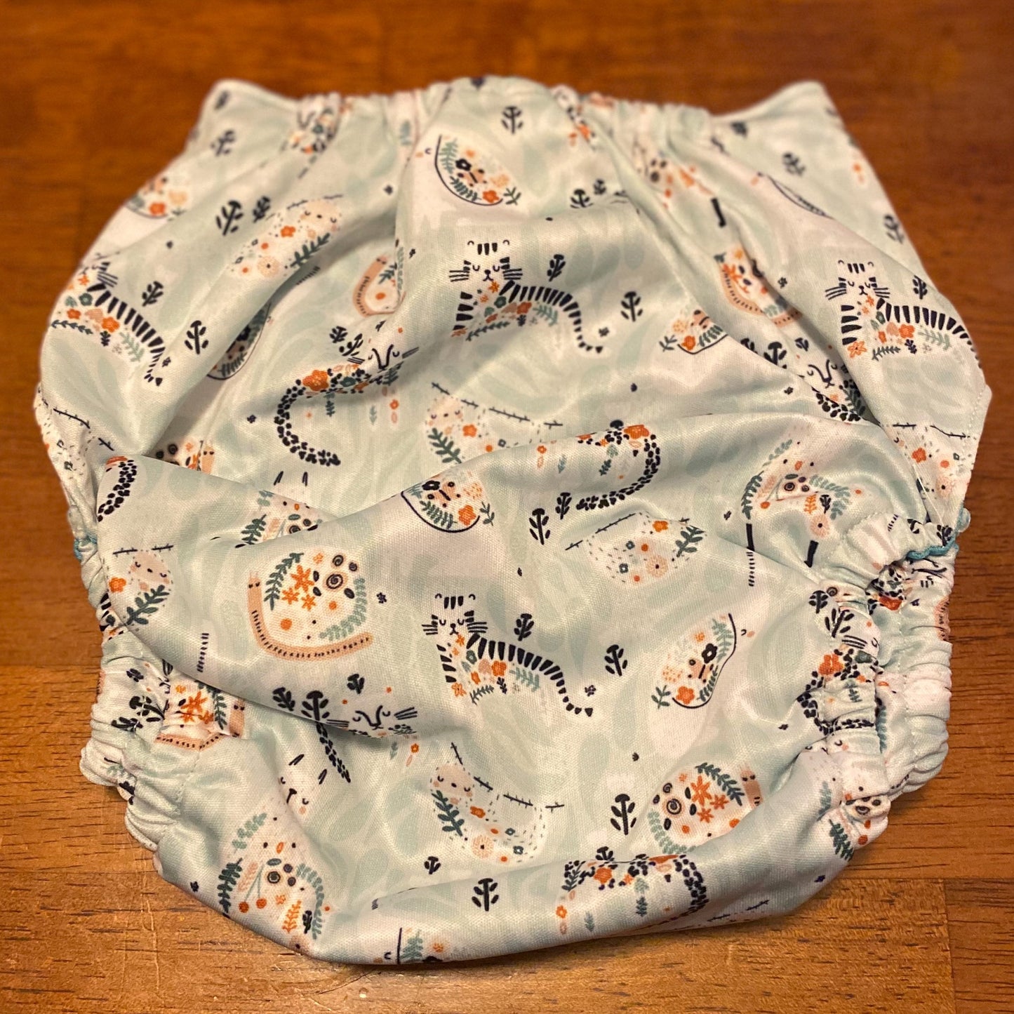 A reusable cloth diaper with blue green lining, elephant, jaguar, sloth, llama and snail folk art print on the outer PUL. Light Gray snaps.